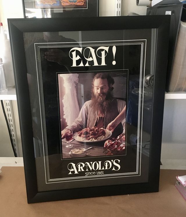 A vintage EAT! Arnold's poster of a bearded man at a table, in a black frame with layered mats