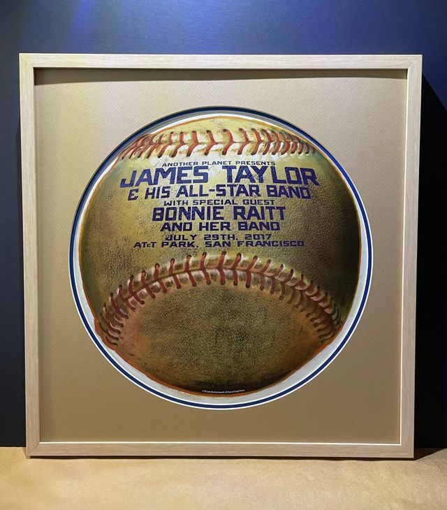 A round baseball-style James Taylor and Bonnie Raitt concert poster showing through a circular mat opening in a white frame