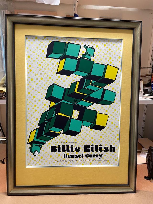 A Billie Eilish gig poster with stacked green cubes, in a silver frame with a gold mat