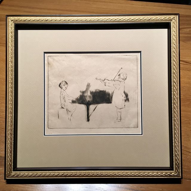 An etching of a pianist and a violinist in an ornate gold frame with a wide cream mat and black inner border