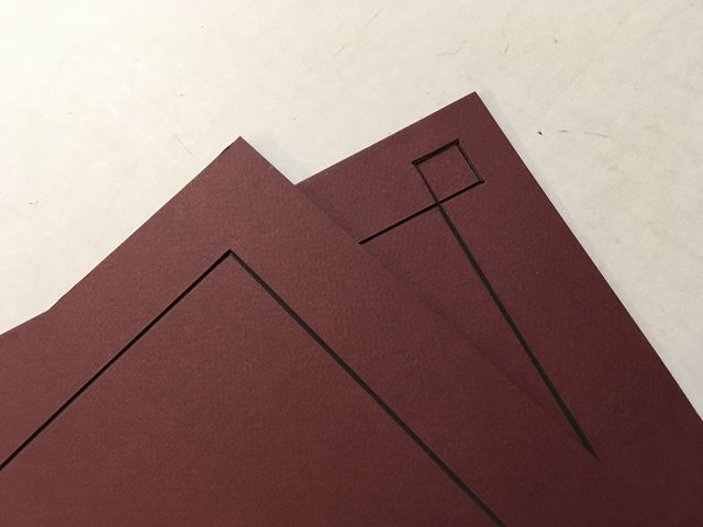 Corners of two burgundy mat boards with cut window openings, one with a small square opening