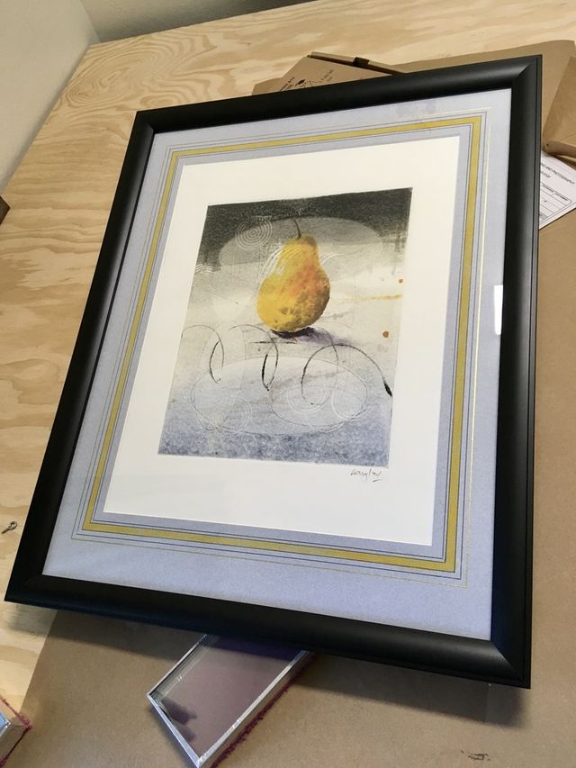 A print of a single pear in a black frame with layered gray, gold and white mats