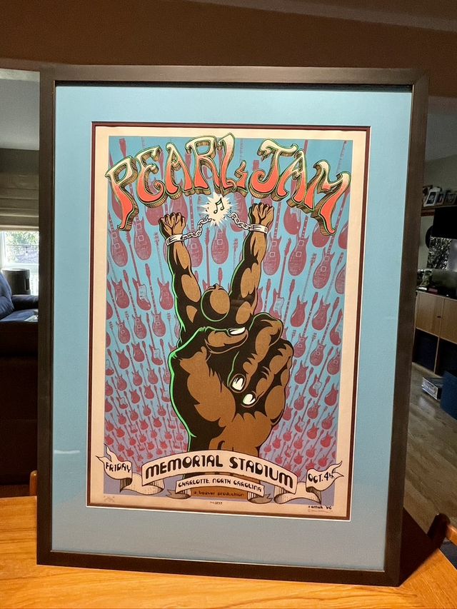 A Pearl Jam concert poster with a raised hand illustration, in a black frame with a light blue mat