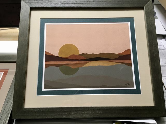 A minimal sunset landscape print in a dark gray frame with teal and cream mats
