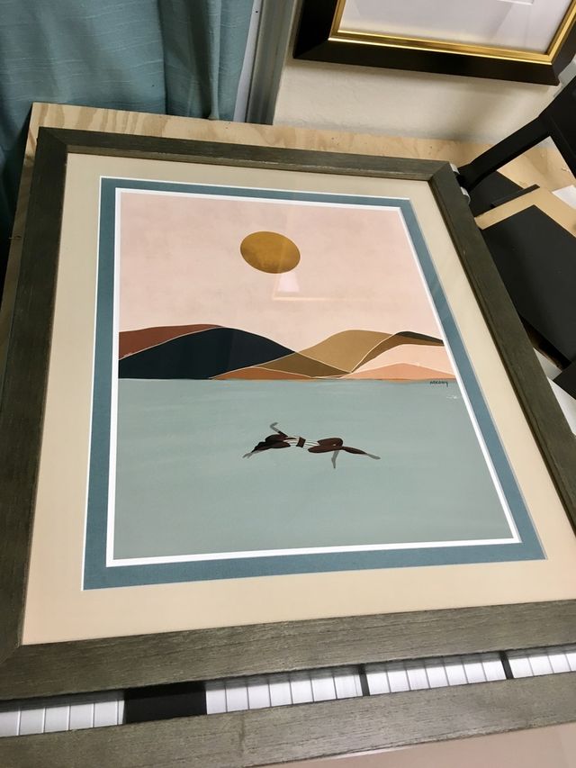 A print of a swimmer in pale water beneath a sun and dunes, in a gray frame with teal and cream mats