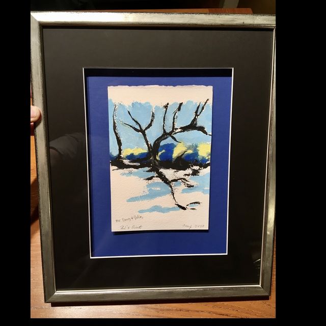 A small painting of a bare tree in snow, in a silver frame with black and blue mats