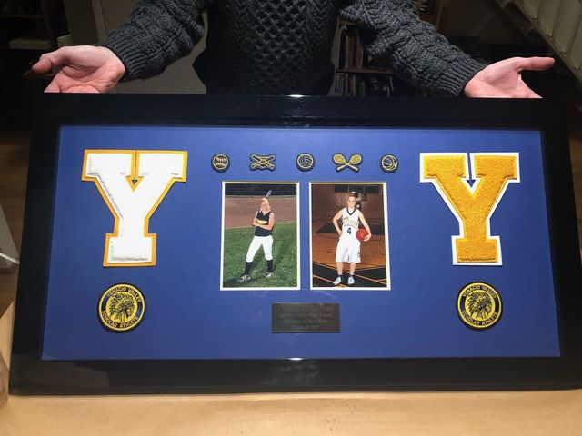 Two varsity letters, two photos, pins and patches arranged on a royal blue mat in a black frame with a small nameplate