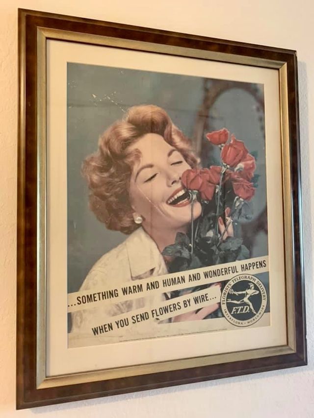 A vintage flowers-by-wire advertisement showing a woman smelling red flowers, in a wood frame with a cream mat